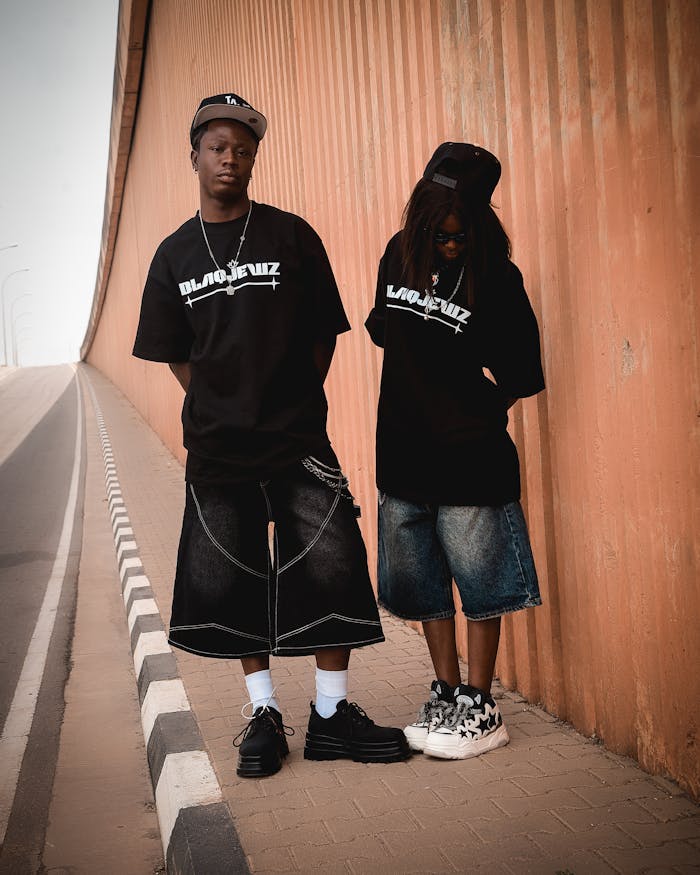 services-03 Two teens in streetwear pose stylishly by an orange wall in an urban setting.