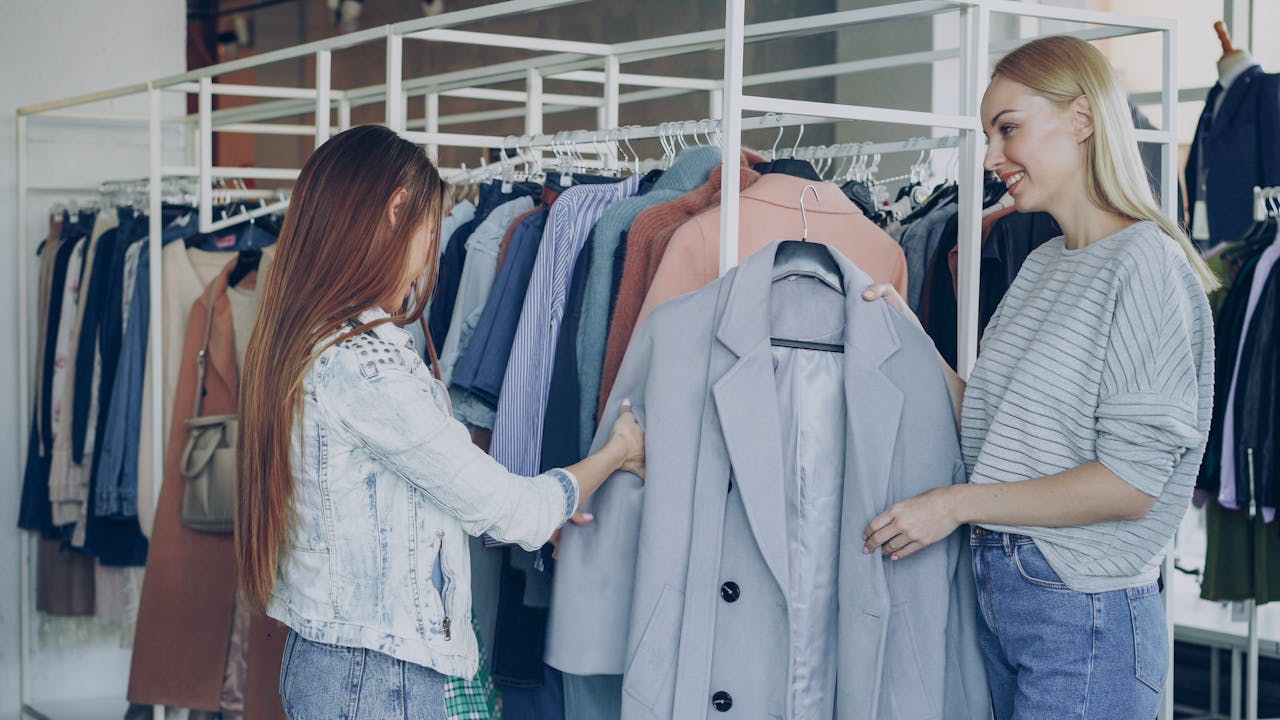 about-02 Two women shopping for jackets in a stylish clothing store, examining coats on a rack.